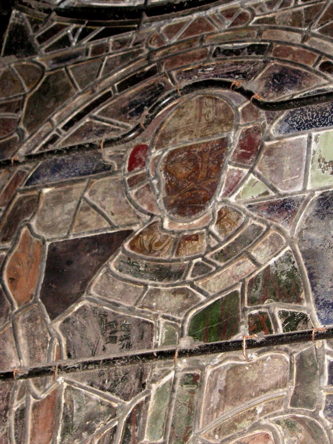 A close view of an old stained-glass panel lit from behind, a haloed figure in grey and dark glass, every piece held by a grid of grey lead strips with bright soldered joins.